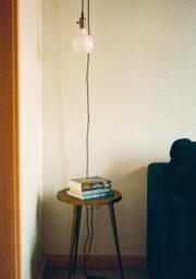 A small wooden side table with a stack of three books stands next to a dark sofa in a room with light-colored walls. A single light bulb hangs above the table with a long cord.