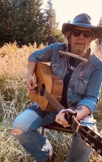 A person wearing a denim outfit, hat, and sunglasses is sitting on a chair outdoors in a field. They are playing an acoustic guitar and have a harmonica holder around their neck. Tall grass and trees are visible in the background.