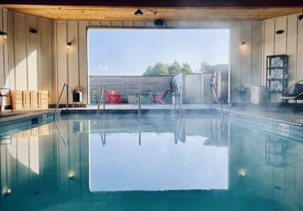 Indoor pool with blue water under a wooden ceiling. Steam rises from the water. Poolside seating includes red chairs outside a large window. The window opens to a patio area visible in the background with more red chairs and some greenery.