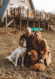 A man and a woman are sitting on the grass with two dogs in front of an A-frame house. The man is petting one dog, while the woman, wearing a coat, is petting the other dog. The yard appears to be spacious with some trees and plants in the background.