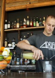 A bartender is pouring a yellow liquid into a glass behind a bar. There is a basket of assorted citrus fruits on the counter. The shelves behind him hold various bottles of alcohol. He is wearing a grey t-shirt with "ADRIFT HOTEL + SPAS" printed on it.